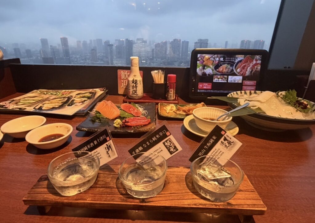 北の味紀行と地酒 北海道 カレッタ汐留店で楽しんだ海鮮料理と北の地酒飲み比べセット。窓の外には東京の景色が広がっている。